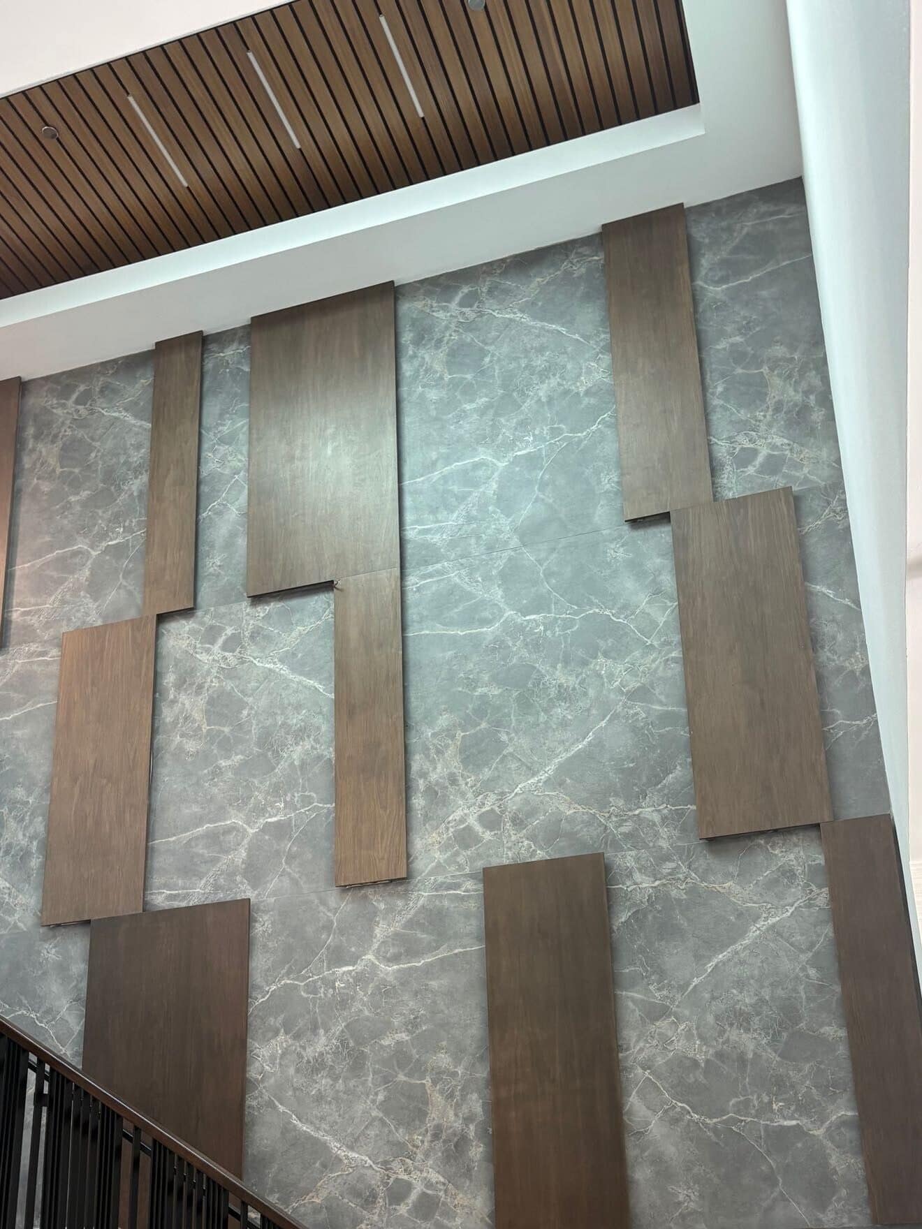 Two-storey commercial lobby wall clad in grey veined stone slab with offset walnut panels and a slatted wood ceiling